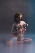 Woman in strappy bridal playsuit and underwear holding flowers against a gradient background