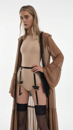 A model wearing a beige bodysuit with black garter straps and thigh-high stockings, styled with a sheer brown fringed robe over a plain white studio background.