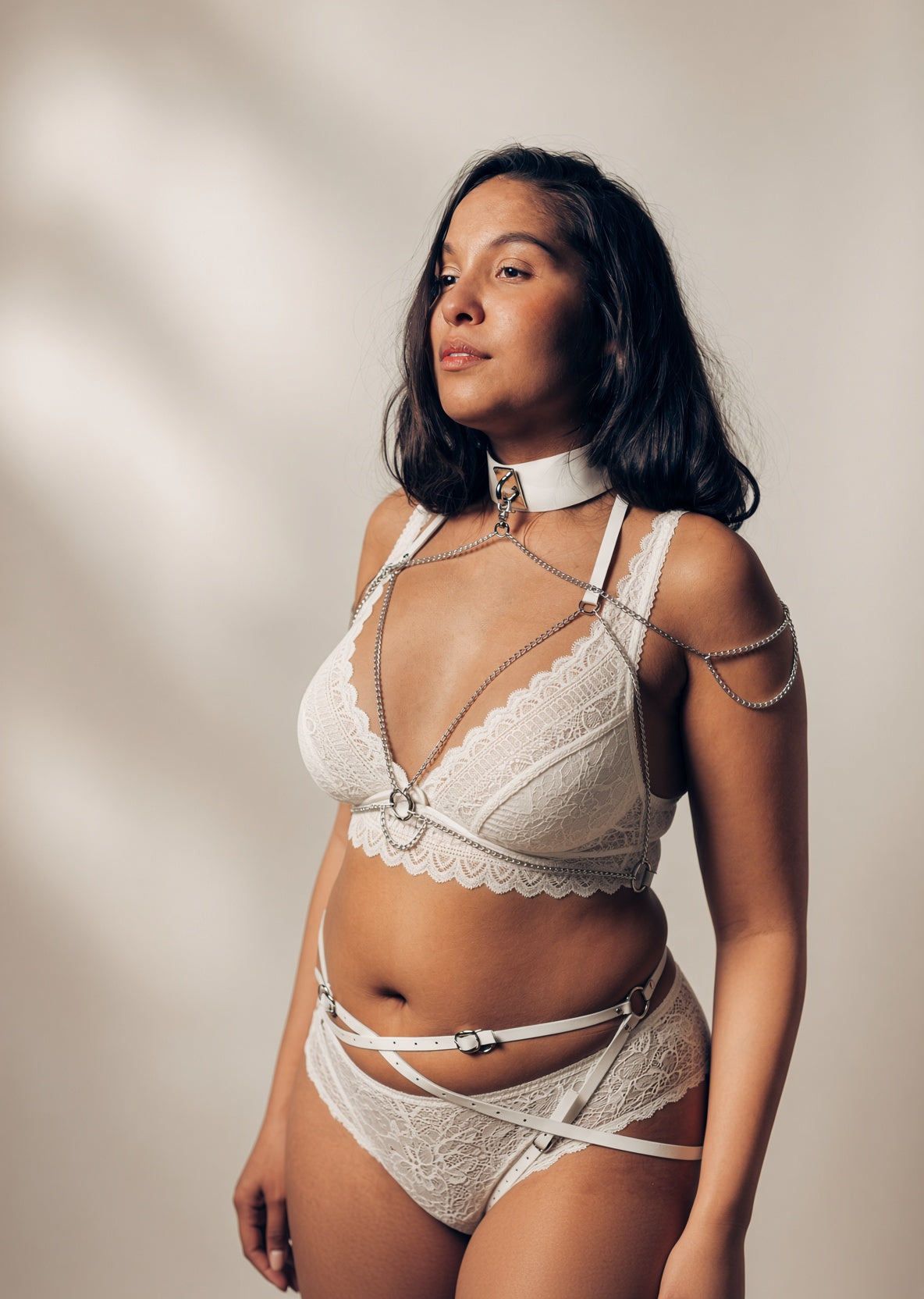 A woman modeling a white lace lingerie set with silver chain harness details, a matching white collar, and strappy waist ties against a warm beige background with soft leaf shadows.