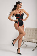 A woman models a black strappy lingerie bodysuit with red trim and garter-style ties, standing in a studio next to a light beige tufted chair.