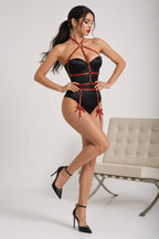 A woman models a black strappy lingerie bodysuit with red trim and garter-style ties, standing in a studio next to a light beige tufted chair.