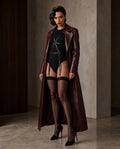 A model stands in a studio setting wearing a long burgundy faux-leather coat over a black bodysuit with a harness-style strap design, black thigh-high stockings, and black heels.