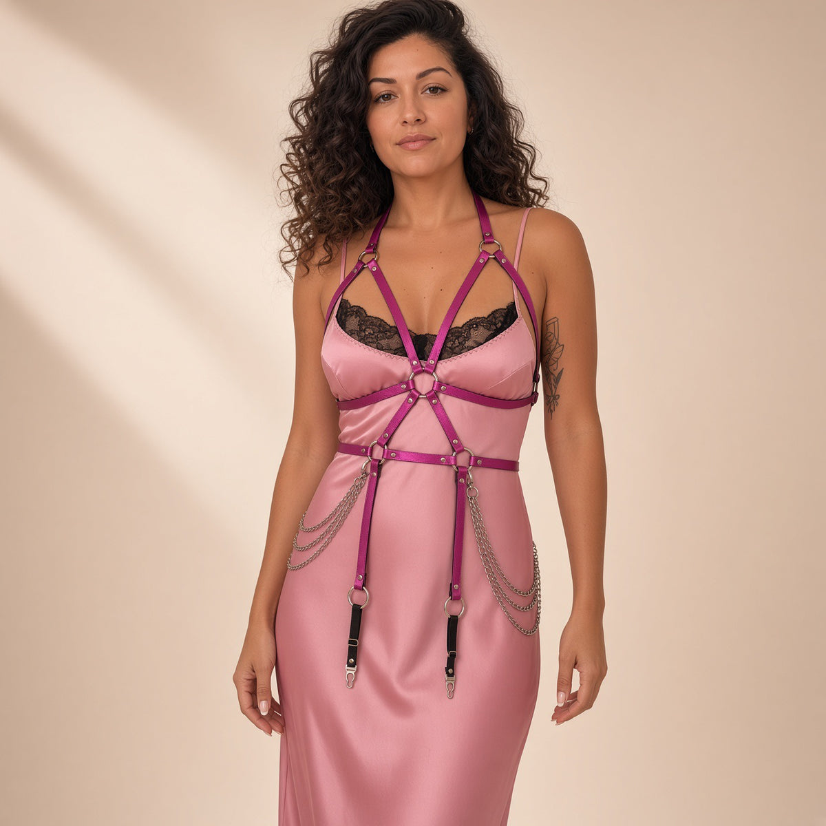 A woman modeling a pink satin slip dress with magenta leather harness-style straps, black lace underwire, and silver chain details against a warm beige studio background.