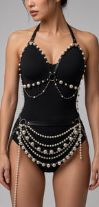 A woman modeling a black one-piece swimsuit with decorative pearl and silver bead chain accents, shown against a plain gray studio background.