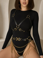 A model wearing a black long-sleeve bodysuit with black faux-leather harness straps and gold-tone metal rings, paired with matching gold chain drapes at the hips, set against a warm beige architectural background.