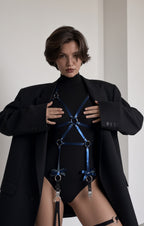 A model wearing a black turtleneck bodysuit with a blue metallic strappy harness and silver O-ring details, styled with an oversized black blazer.