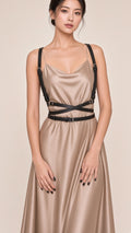A woman modeling a satin-like sleeveless dress in a warm taupe-beige color with black suspenders and a black waist belt, photographed against a plain light beige studio background.