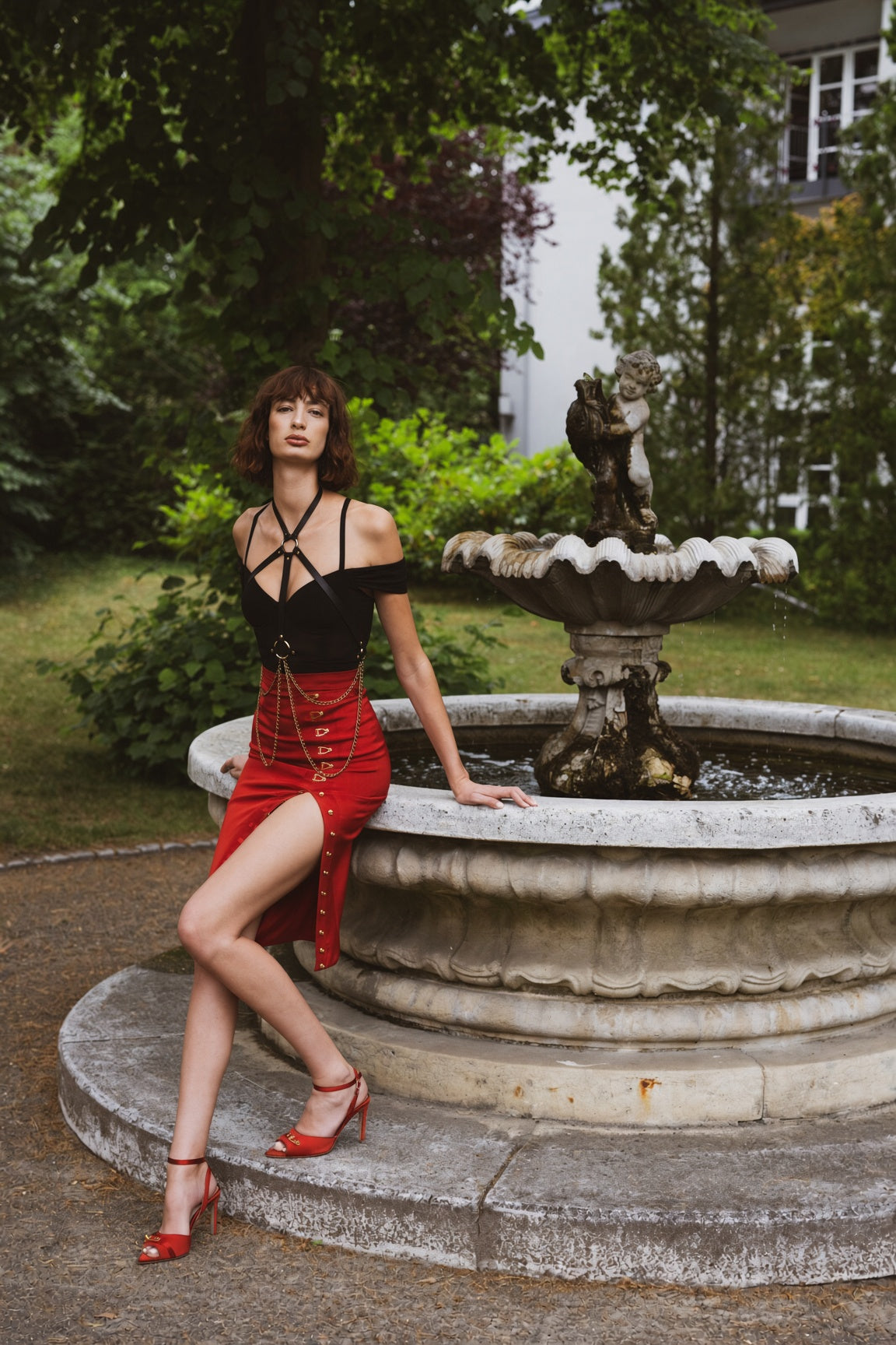 Woman in a black top and red skirt standing by a fountain in a garden.