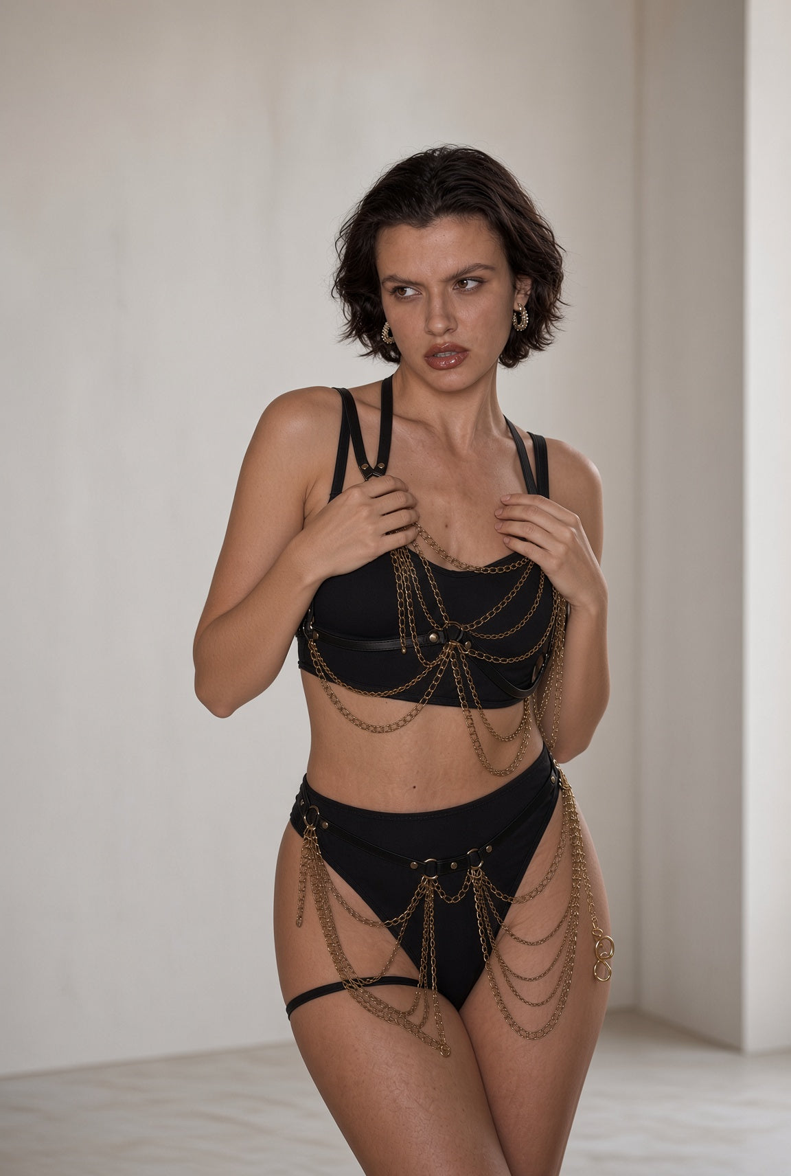 A model wearing a black lingerie set with elaborate gold chain draping, photographed in a minimalist studio setting with a warm off-white background.