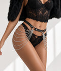 A close-up of a model wearing black lace lingerie with a black faux-leather waist belt and silver chain drapes, styled with a black feathered jacket against a bright white background.