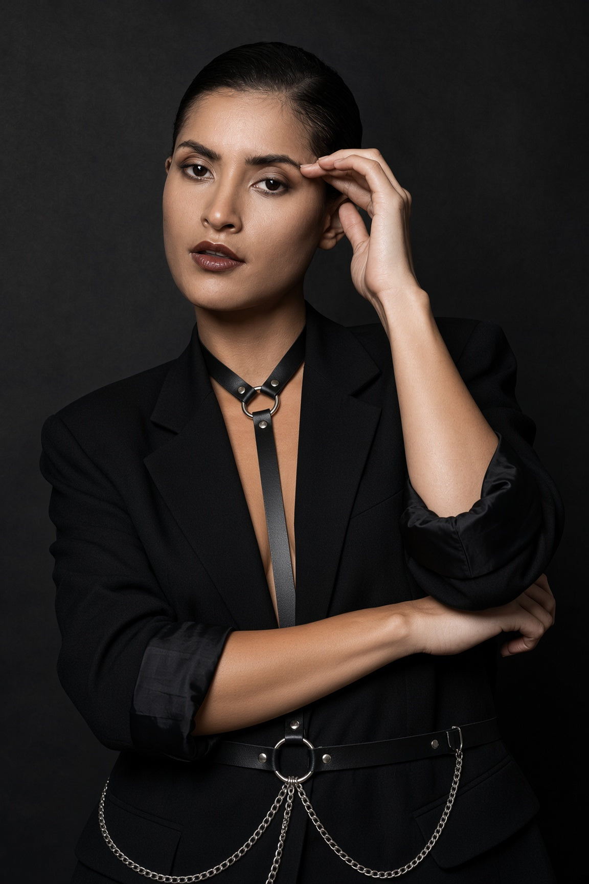 A woman with slicked-back dark hair models a black leather choker collar with a large silver ring, styled with a black blazer and a black belt with silver chain details against a dark studio background.